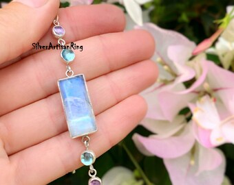 May include: A silver chain bracelet featuring a large rectangular moonstone pendant and smaller round blue and purple gemstones. The bracelet is held in a hand against a backdrop of pink flowers.