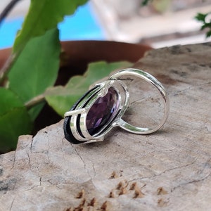May include: A silver ring with a large, oval-shaped purple gemstone set in a silver band. The band has three silver lines running down the side. The ring is on a wooden surface.