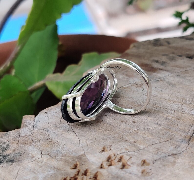 May include: A silver ring with a large, oval-shaped purple gemstone set in a silver band. The band has three silver lines running down the side. The ring is on a wooden surface.
