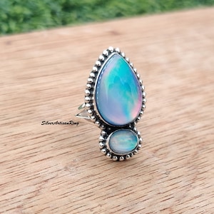 May include: A silver ring with two iridescent opal stones. The larger stone is teardrop shaped and the smaller stone is oval. The ring is set with small silver beads around the stones.