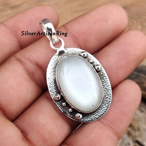 Op de afbeelding: Een zilveren hanger met een witte ovale edelsteen gezet in een getextureerd ovaal frame. De hanger heeft een lus om aan een ketting te bevestigen.