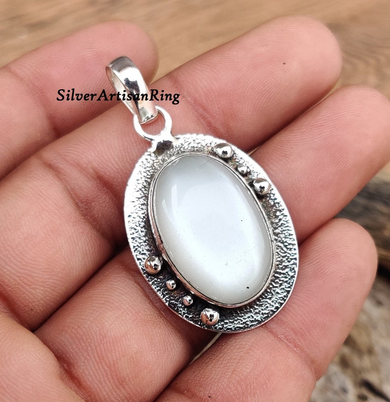 Op de afbeelding: Een zilveren hanger met een witte ovale edelsteen gezet in een getextureerd ovaal frame. De hanger heeft een lus om aan een ketting te bevestigen.