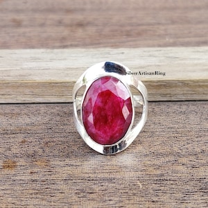 Könnte beinhalten: Ein silberner Ring mit einem großen, ovalen, roten Edelstein, der in einem filigranen, durchbrochenen Design gefasst ist. Der Ring liegt auf einer Holzoberfläche. SilverArtisanRing