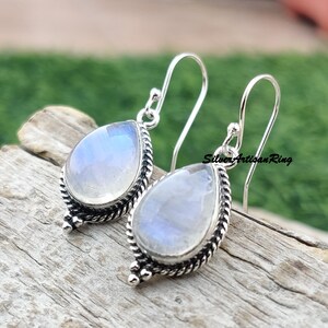 May include: A pair of silver dangle earrings with teardrop-shaped moonstone gemstones. The earrings have a delicate, intricate design with a twisted rope border and small silver beads.