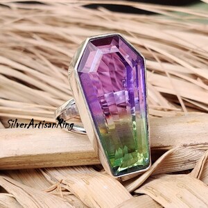 May include: A silver ring featuring a large, faceted, coffin-shaped gemstone. The gemstone is a gradient of purple, blue, and green.