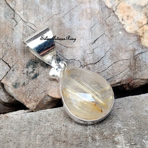 May include: A silver pendant with a teardrop-shaped, clear quartz stone containing golden, hair-like inclusions. The pendant has a silver bail and is set against a rustic wooden background. The text "Silver Artisan Ring" is visible on the image.