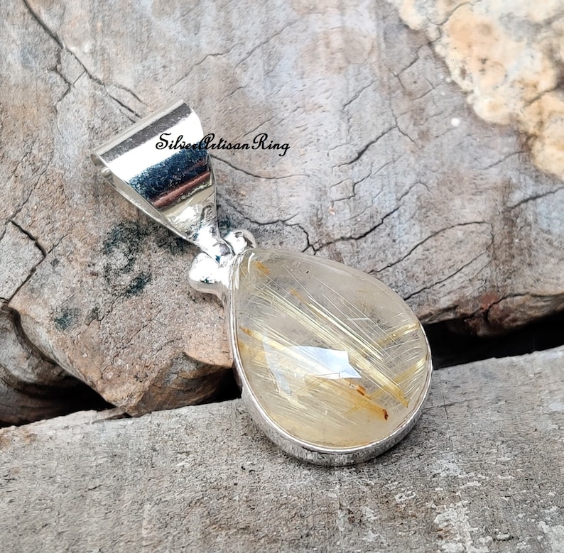 May include: A silver pendant with a teardrop-shaped, clear quartz stone containing golden, hair-like inclusions. The pendant has a silver bail and is set against a rustic wooden background. The text "Silver Artisan Ring" is visible on the image.