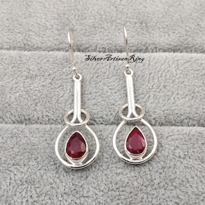 Könnte beinhalten: Silberne Ohrringe mit einem tropfenförmigen, tiefroten Edelstein, der in einen kreisförmigen Rahmen gefasst ist. Die Ohrringe haben ein langes, vertikales Design mit einem Haken für durchbohrte Ohren. Der Text "SilverArtisanRing" ist sichtbar.