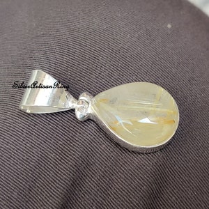May include: A teardrop-shaped pendant with a silver bail and setting. The pendant features a translucent, light-yellow stone with golden inclusions. The pendant is set against a dark gray background.