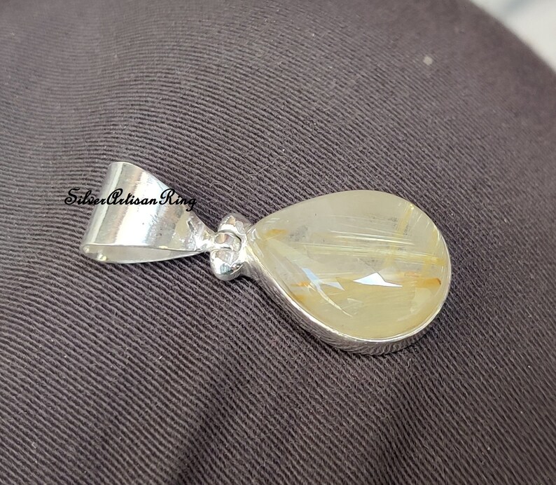 May include: A teardrop-shaped pendant with a silver bail and setting. The pendant features a translucent, light-yellow stone with golden inclusions. The pendant is set against a dark gray background.