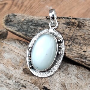 Op de afbeelding: Een zilveren hanger met een witte ovale edelsteen in een getextureerd ovaal frame. Het frame heeft kleine zilveren kralen rond de rand.