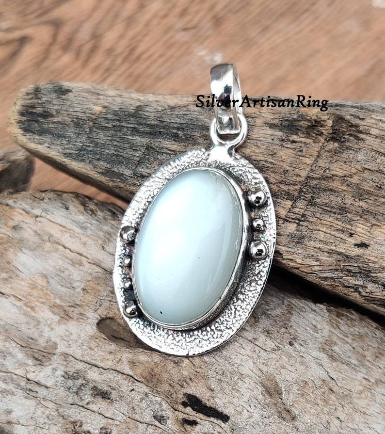 Op de afbeelding: Een zilveren hanger met een witte ovale edelsteen in een getextureerd ovaal frame. Het frame heeft kleine zilveren kralen rond de rand.