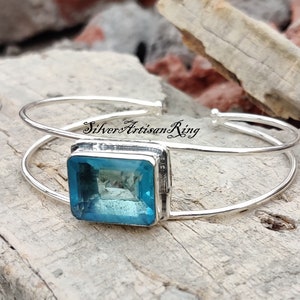 Op de afbeelding: Een zilveren armband met een grote, rechthoekige, lichtblauwe edelsteen die in een zilveren zetting is gezet. De armband is open en heeft een eenvoudig ontwerp.