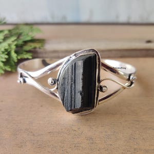 Könnte beinhalten: Ein silberfarbenes Armreif-Armband mit einem großen, rechteckigen schwarzen Turmalinstein. Das Armband hat ein geschwungenes Design mit dekorativen Wirbeln und kleinen runden Akzenten. Der Stein ist in einer Lünettenfassung gefasst.