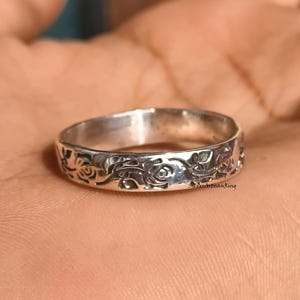 May include: A silver ring with a floral design. The ring features a continuous pattern of roses and leaves around the band. The ring is shown on a hand.