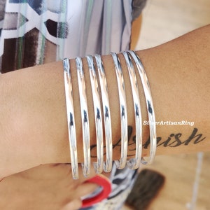 May include: Seven polished silver bangles are stacked on an arm. The bangles are smooth and circular, reflecting light. The arm is wearing a striped shirt. The text "SilverArtisanRing" and a tattoo are visible.