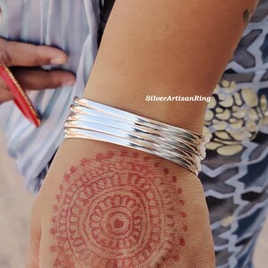 May include: A collection of polished silver bangles stacked on a wrist. The bangles are reflective and catch the light. The hand displays a henna pattern. The image is taken outside.