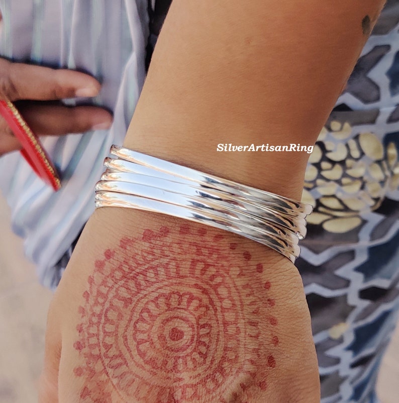 May include: A collection of polished silver bangles stacked on a wrist. The bangles are reflective and catch the light. The hand displays a henna pattern. The image is taken outside.
