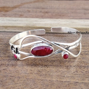 Könnte beinhalten: Ein silbernes Armreif-Armband mit einem roten Edelstein und zwei kleineren roten Edelsteinen. Das Armband hat ein zartes, filigranes Design.