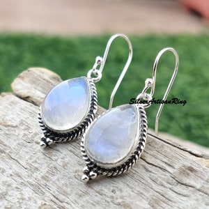 May include: A pair of silver dangle earrings with teardrop-shaped moonstone gemstones. The earrings have a delicate, intricate design with a twisted rope border and small silver beads.