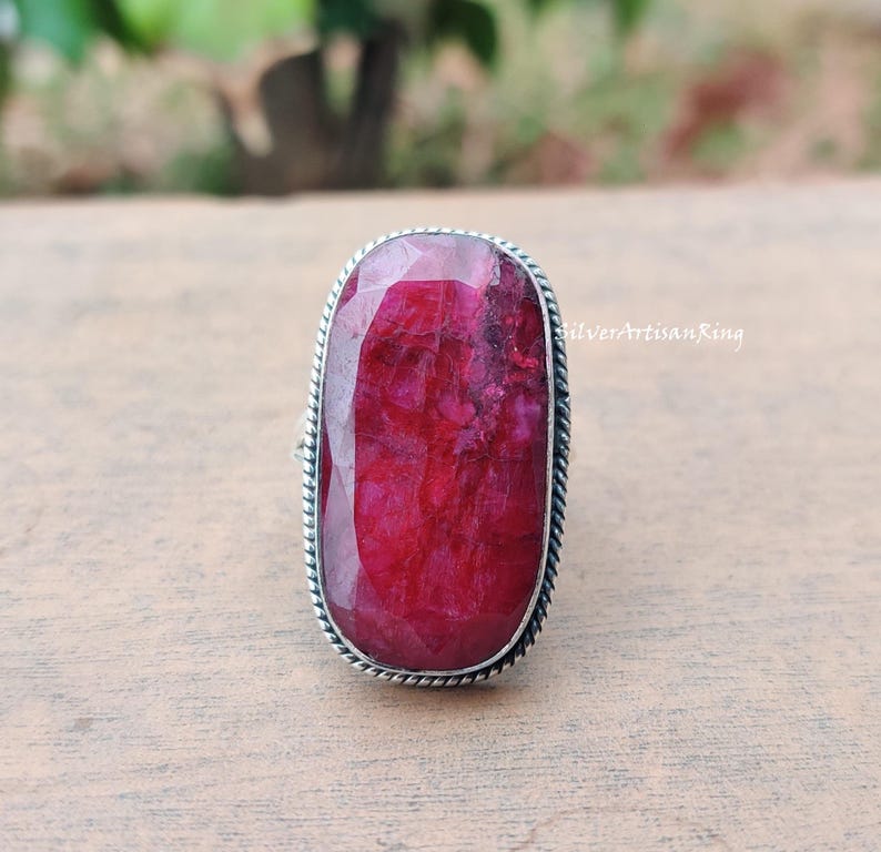 May include: A silver ring featuring a large, rectangular ruby gemstone. The ruby is a rich red hue, set within a silver band with a twisted rope border. The ring is presented on a wooden surface.