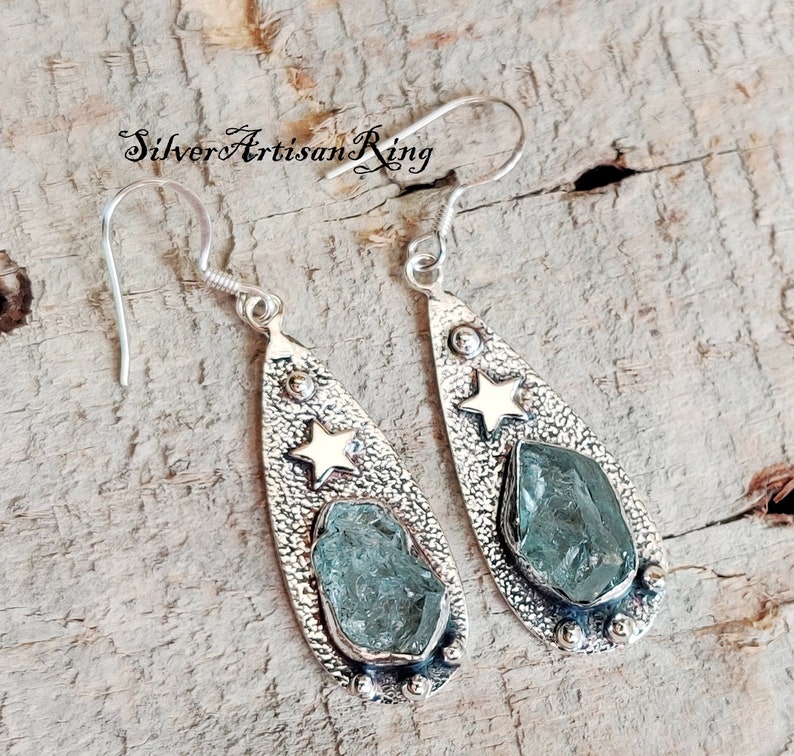 May include: A pair of silver teardrop-shaped earrings with a textured finish. Each earring features a light blue gemstone and a gold star.