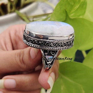 May include: A silver ring with an oval-shaped moonstone set in a filigree design. The ring has a textured band and a beaded edge.