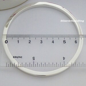 May include: A polished silver bangle with a square profile. A ruler in inches measures the bracelet, showing a diameter of approximately 6.35 cm. The bracelet is displayed on a light-colored surface.