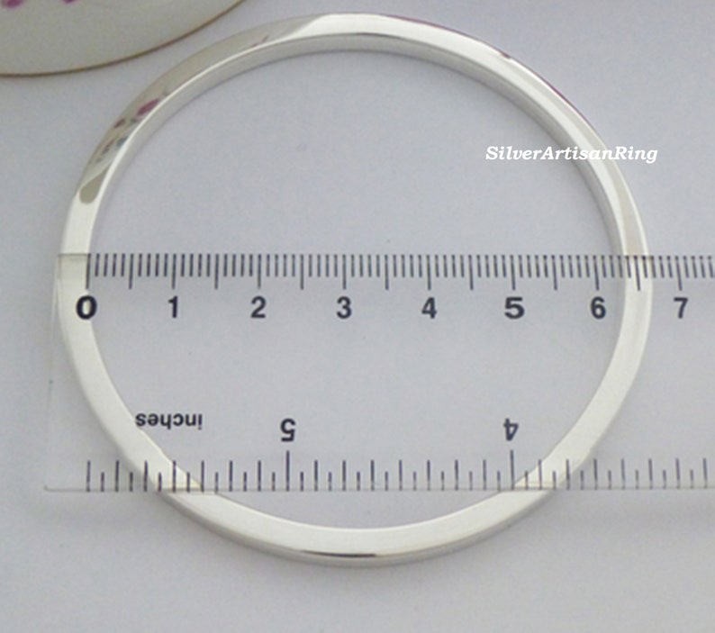 May include: A polished silver bangle with a square profile. A ruler in inches measures the bracelet, showing a diameter of approximately 6.35 cm. The bracelet is displayed on a light-colored surface.