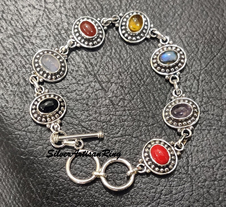 May include: Silver bracelet with seven oval-shaped gemstones set in silver bezels. The gemstones are red, yellow, blue, black, purple, and two shades of blue. The bracelet has a toggle clasp.