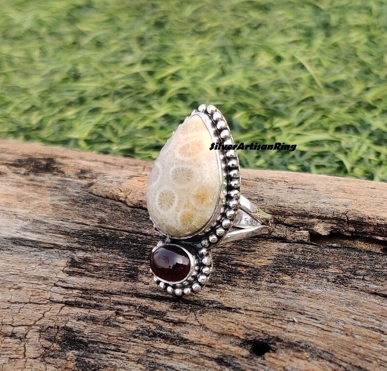 Op de afbeelding: Een zilveren ring met een traanvormige versteende koraalsteen en een kleinere rode edelsteen. De ring is bezet met kleine zilveren kralen rond de stenen.