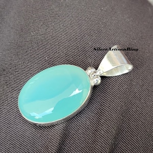 May include: Oval-shaped pendant with a light blue gemstone set in a silver metal frame. The pendant has a polished, glossy surface and a simple bail for a chain. The jewelry is displayed on a dark fabric background.
