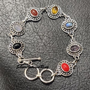 May include: Silver bracelet with seven oval-shaped gemstones set in silver bezels. The gemstones are red, orange, blue, black, purple, and two shades of blue. The bracelet has a toggle clasp.