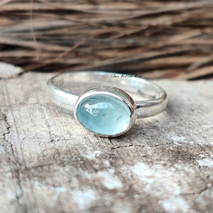 Puede incluir: Un anillo de plata con una piedra ovalada azul claro. El anillo est&aacute; sobre una superficie de madera. "Silver Artisan Ring" est&aacute; escrito en el anillo.