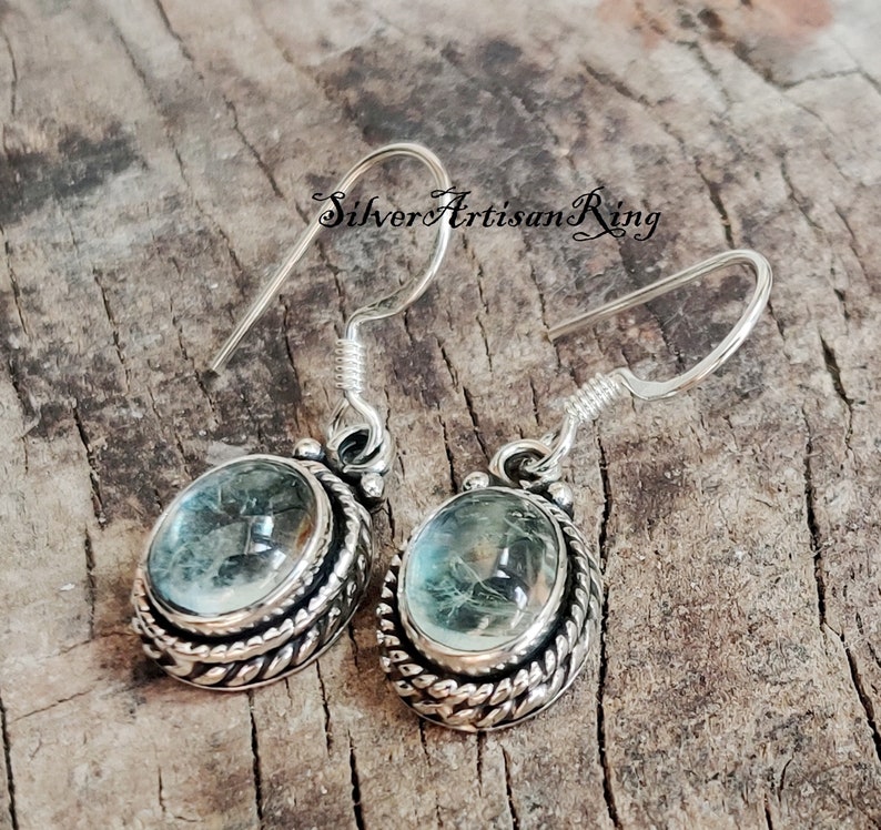 May include: Silver earrings with a light blue oval gemstone set in a silver bezel with a rope-like design. The earrings have a hook closure.