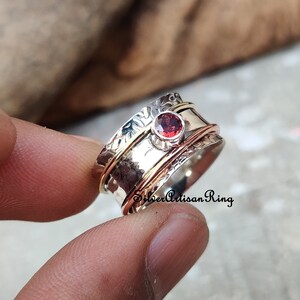May include: A silver spinning ring with a red gemstone and an engraved floral design. The ring has a copper band and is made by Silver Artisan Ring.