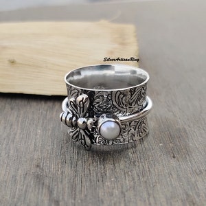 Könnte beinhalten: Ein silberner Drehring mit Libellen-Design und einer weißen Perle. Der Ring ist mit einem floralen Muster strukturiert.