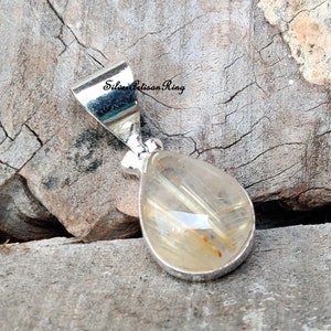 May include: A teardrop-shaped pendant with a silver setting. The pendant features a clear, golden-colored stone with internal golden-brown inclusions. The pendant is displayed on a weathered wooden surface.