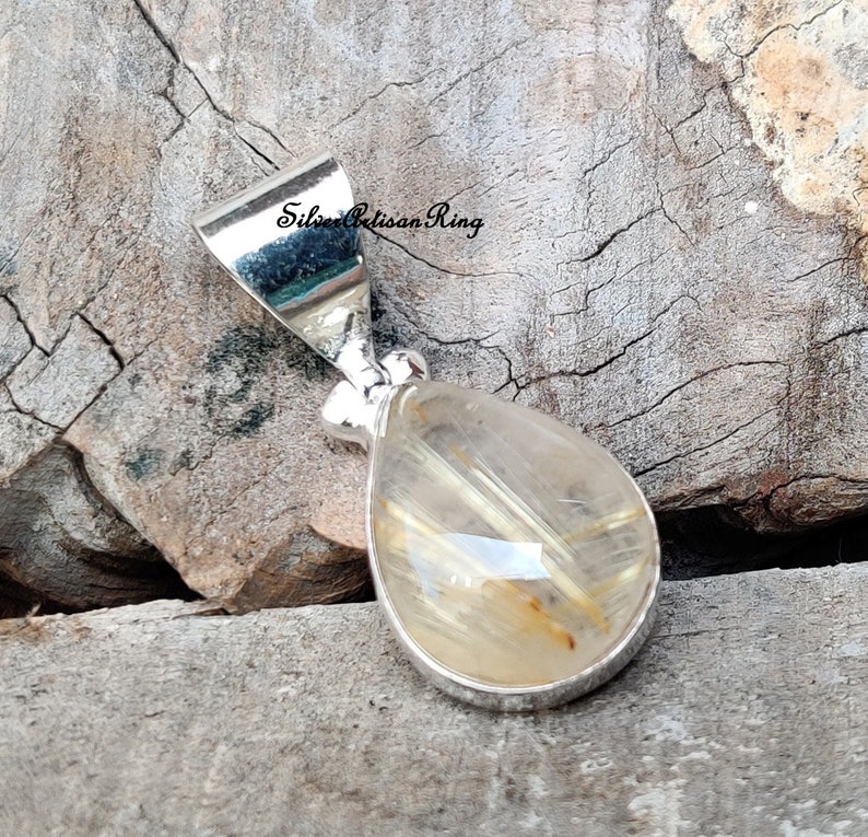 May include: A teardrop-shaped pendant with a silver setting. The pendant features a clear, golden-colored stone with internal golden-brown inclusions. The pendant is displayed on a weathered wooden surface.