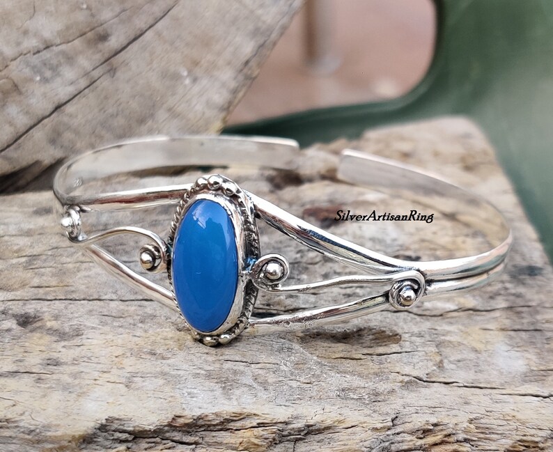 K&ouml;nnte beinhalten: Ein silbernes Armreif-Armband mit einem blauen ovalen Edelstein, der in einer silbernen Fassung sitzt. Das Armband hat ein zartes, verziertes Design mit drei silbernen B&auml;ndern und kleinen silbernen Perlen.