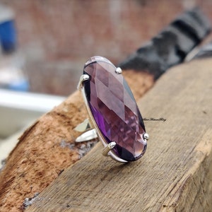 May include: A silver ring with a large, faceted, oval-shaped amethyst gemstone. The ring is set on a wooden surface.