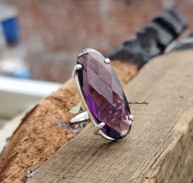 May include: A silver ring with a large, faceted, oval-shaped amethyst gemstone. The ring is set on a wooden surface.