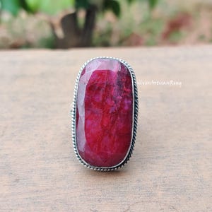 May include: A silver ring featuring a large, rectangular ruby gemstone. The ruby is a rich red hue, set within a silver band with a twisted rope border. The ring is presented on a wooden surface.