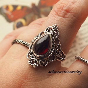 Könnte beinhalten: Ein silberner Ring mit einem verzierten Design zeigt einen tropfenförmigen roten Edelstein. Der Ring wird an einem Finger getragen.