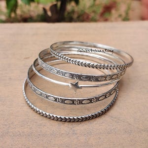 Könnte beinhalten: Ein Stapel silberner Armreifen mit aufwendigen Designs. Einige Armreifen weisen ein Blattmuster auf, andere florale und ovale Muster. Ein Armreif hat einen Sternanhänger. Der Text "SilverArtisanRing" ist sichtbar. Diese Armbänder sind ideal für einen eleganten Look.