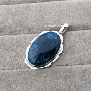 K&ouml;nnte beinhalten: Ein silberner Anh&auml;nger mit einem blauen Edelstein, der in einer gewellten Fassung sitzt. Der Anh&auml;nger ist oval geformt und hat eine &Ouml;se zum Anbringen einer Kette.