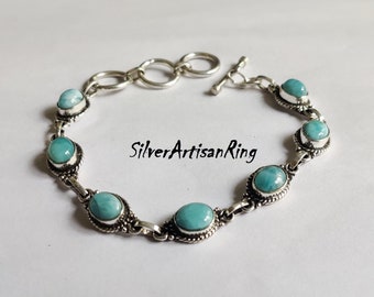 May include: A silver bracelet with a toggle clasp features six oval-shaped blue and white gemstone beads. The bracelet is adorned with intricate silver detailing around each bead. The text "SilverArtisanRing" is visible in the image.