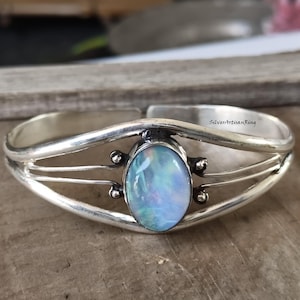 May include: A silver cuff bracelet with an oval, iridescent opal gemstone at the center. The bracelet has a curved design with three parallel bands and small decorative accents. The text "SilverArtisanRing" is visible on the bracelet.
