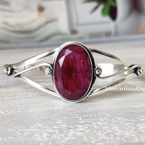 Könnte beinhalten: Ein silbernes Armreif-Armband mit einem großen, ovalen, tief roten Edelstein in der Mitte. Das Armband hat ein zartes, gedrehtes Design.