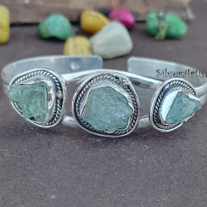 May include: Silver cuff bracelet with three green aquamarine stones set in silver. The bracelet is open and has a simple design.